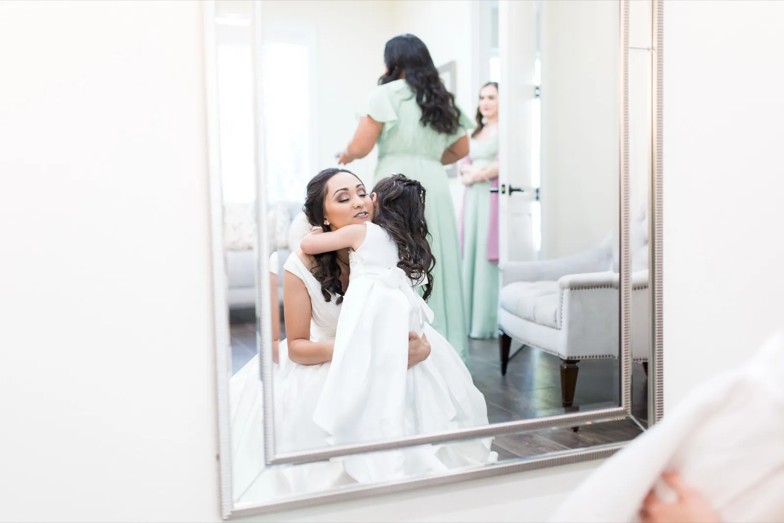 A bride crouches to hug a young girl in a white dress, the moment reflected in a tall mirror