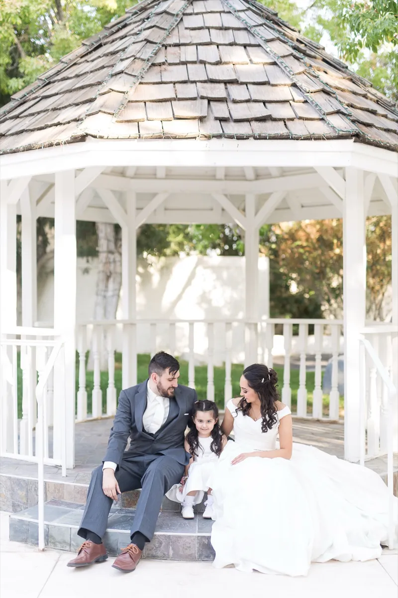 Couple sitting on the steps of the white wooden gazebo with a flower girl between them