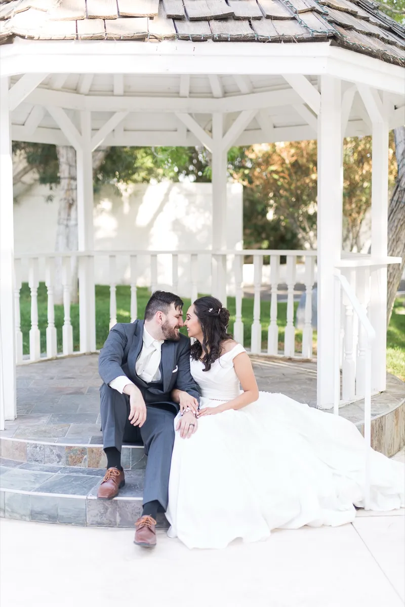 Couple sitting together on the steps of the white wooden gazebo, the bride's skirt spilling down the stairs
