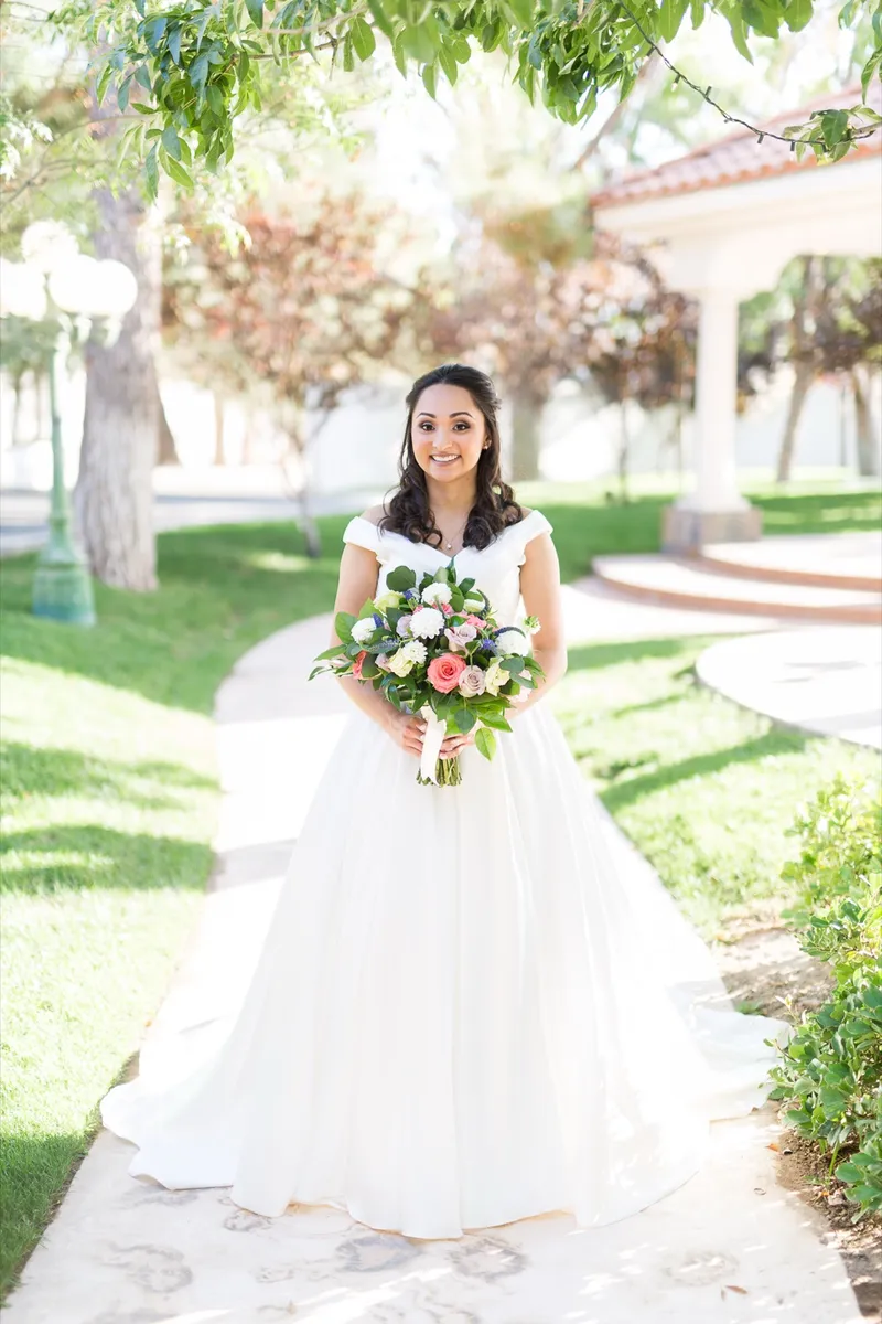 Bride standing on a garden path holding a peach and white bouquet with trees closing in on both sides