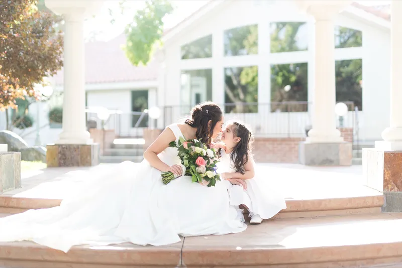 Bride leaning down to kiss a flower girl on the courtyard steps, both of them holding bouquets