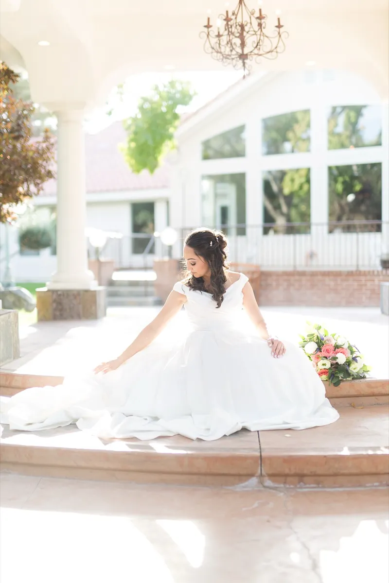 Bride sitting on the edge of a stone fountain with her gown spread out around her and the venue windows behind