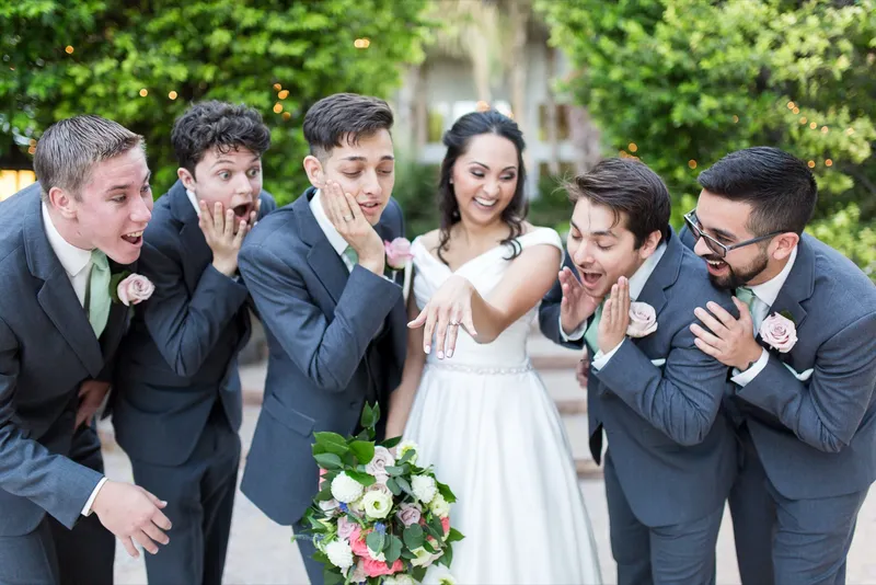 Groomsmen clutching their faces in mock shock at the bride's ring while she holds her hand out