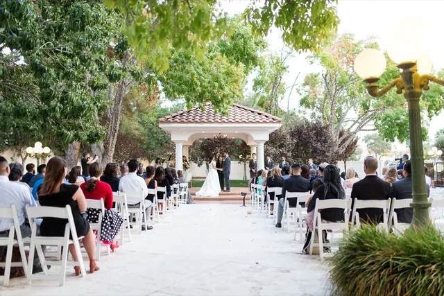 Wide view down the marble aisle to a ceremony under a white columned pavilion with a tile roof, guests seated in white folding chairs on both sides and big trees strung with small lights overhead