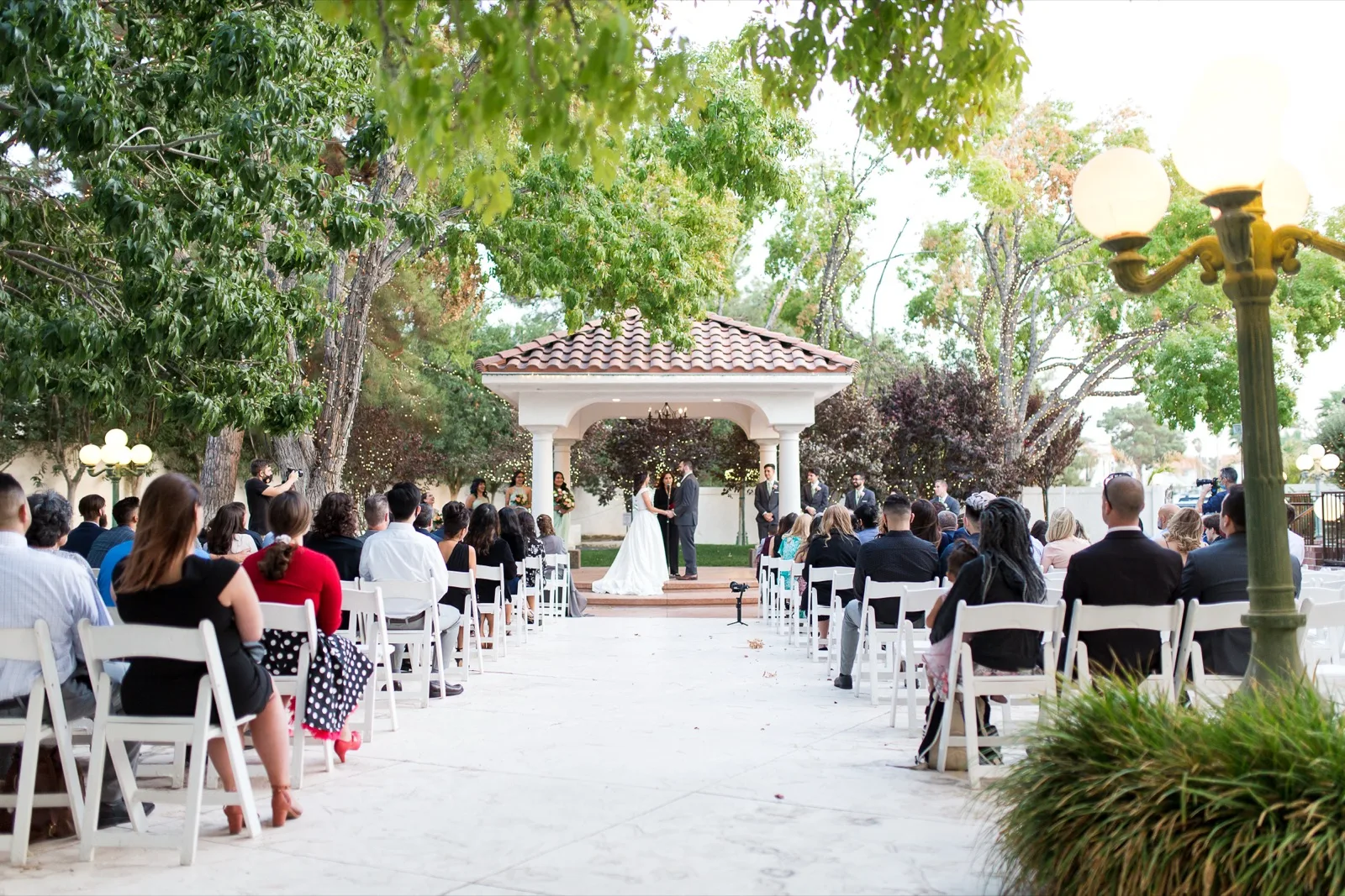 Wide view down the marble aisle to a ceremony under a white columned pavilion with a tile roof, guests seated in white folding chairs on both sides and big trees strung with small lights overhead
