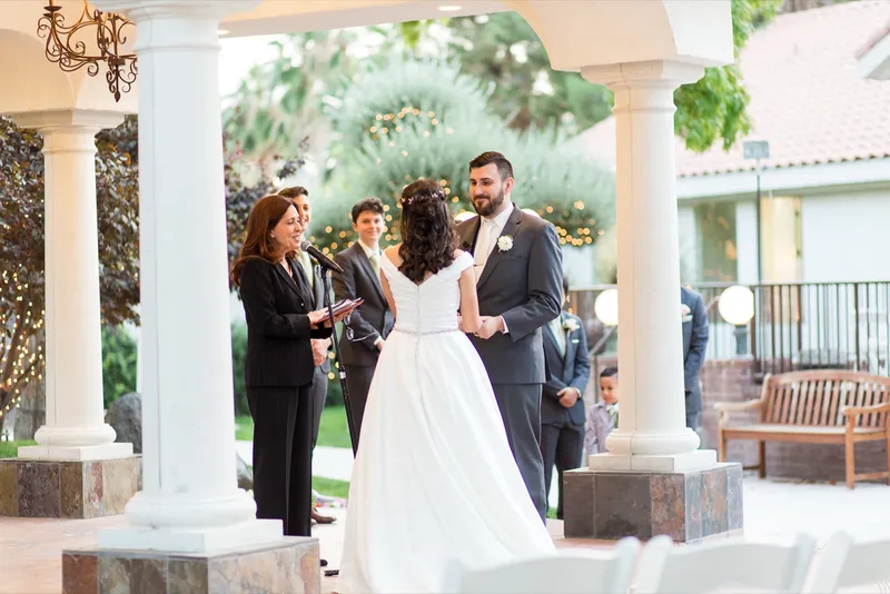 Couple standing between the white pavilion columns with the officiant and a chandelier hanging above them