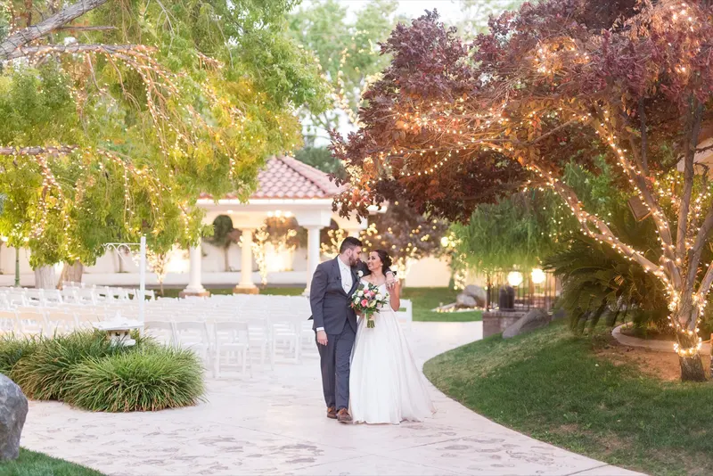 A bride and groom walk a garden path past rows of white ceremony chairs, string lights glowing in the trees