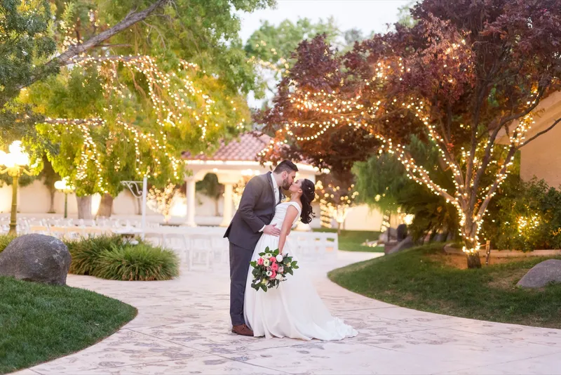 Couple leaning into a kiss under the lit trees, her bouquet held up beside them