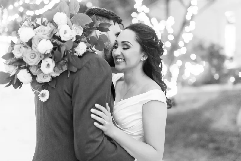 Black and white close portrait of the couple with her bouquet at the edge of the frame and lights out of focus behind