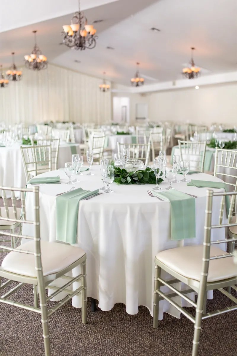 Round reception table with mint napkins and a low greenery runner, chandeliers overhead