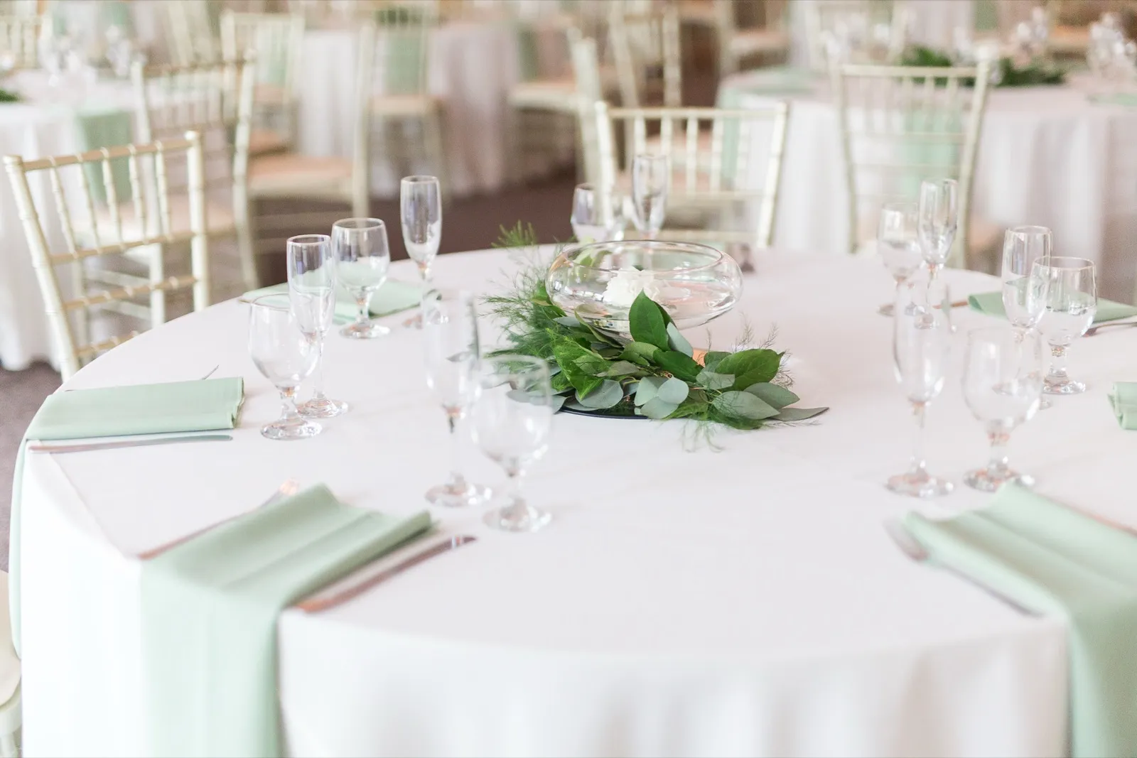 A round table in white linen and mint napkins, set with glassware around a glass bowl of greenery