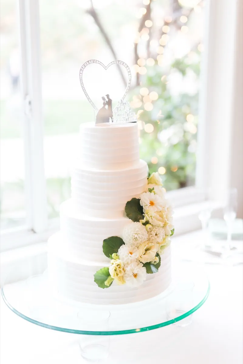 Three tier white cake trimmed with yellow roses and greenery, a heart topper on top, standing in a window