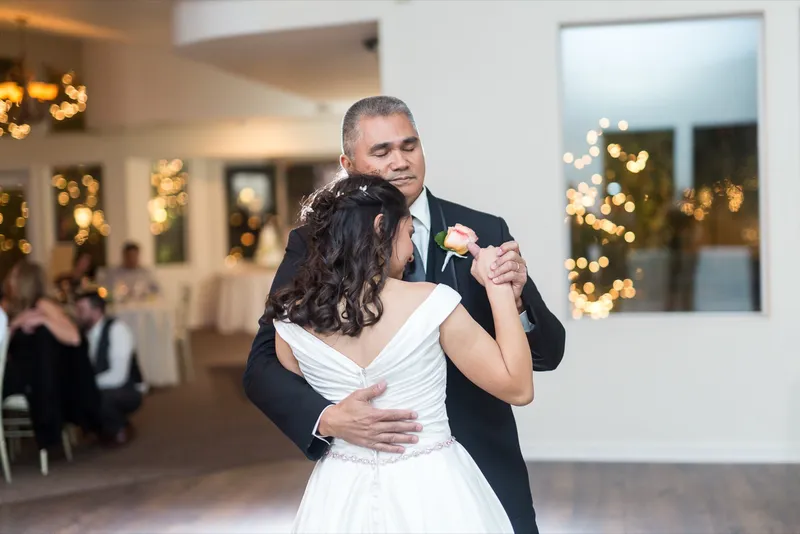 Bride dancing with a parent on the reception floor, string lights showing through the doorway behind