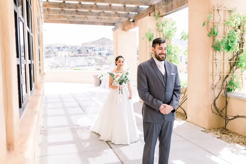 Groom in a grey suit waiting under the wood beams of the covered walkway while his bride comes up behind him