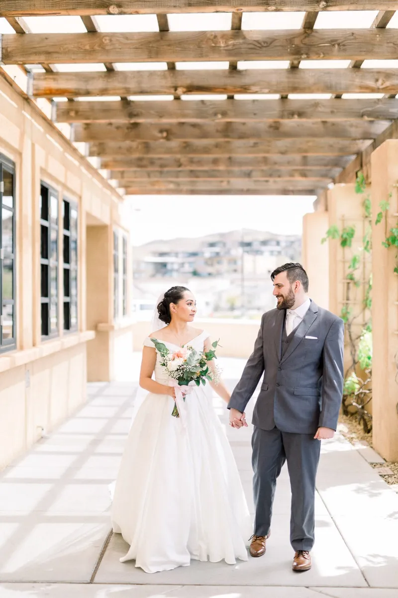 Couple walking hand in hand down the shaded walkway, her train trailing over the pavers