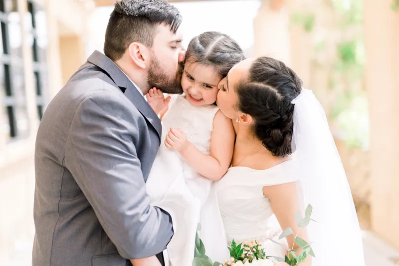 Bride and groom each kissing a little girl in an ivory dress on the cheek while she grins between them