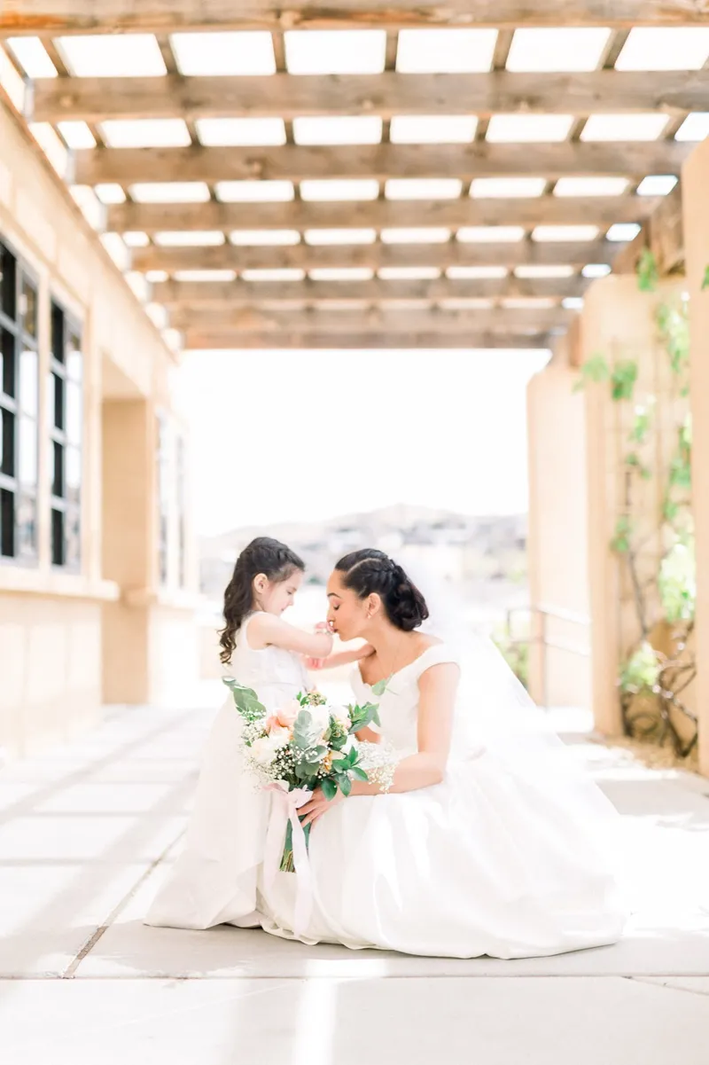 Bride kneeling on the walkway in her full skirt to touch foreheads with a little girl in an ivory dress