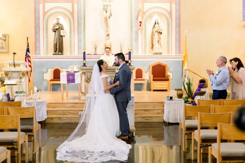 Couple standing together at the altar with her lace veil trailing down the step behind them, the priest and two guests nearby
