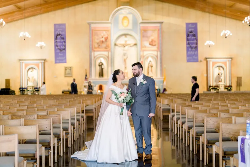 Couple standing in the middle of the church with rows of empty wooden chairs behind them