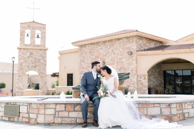 Bride and groom sitting close together on the stone wall of the church fountain, the bell tower and the tile roofed church behind them