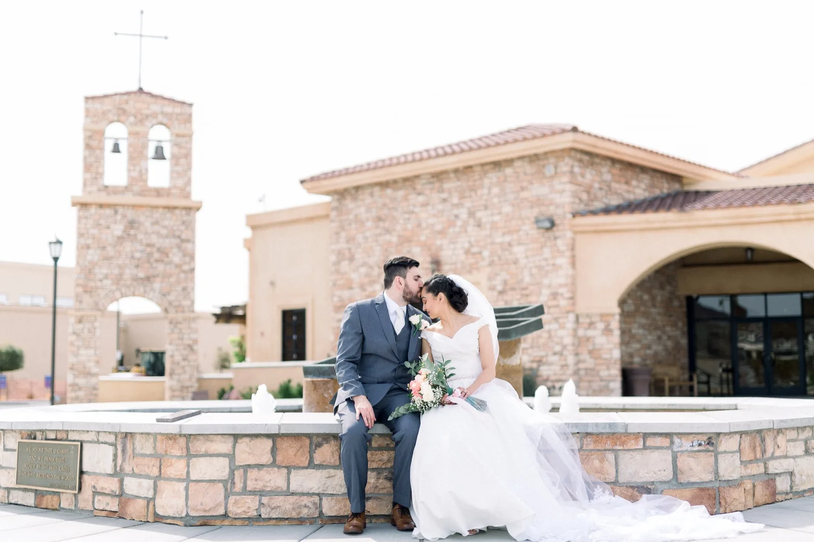 Bride and groom sitting close together on the stone wall of the church fountain, the bell tower and the tile roofed church behind them