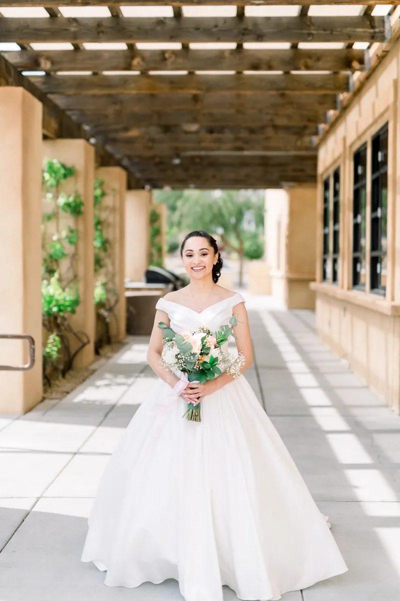 Bride standing under the vine covered walkway holding her peach and white bouquet