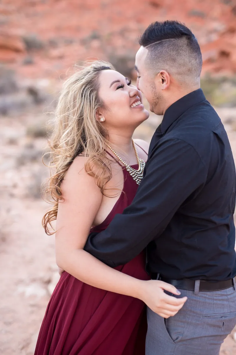 Woman in a burgundy gown laughing with her forehead against her fiance's cheek, red rock behind them