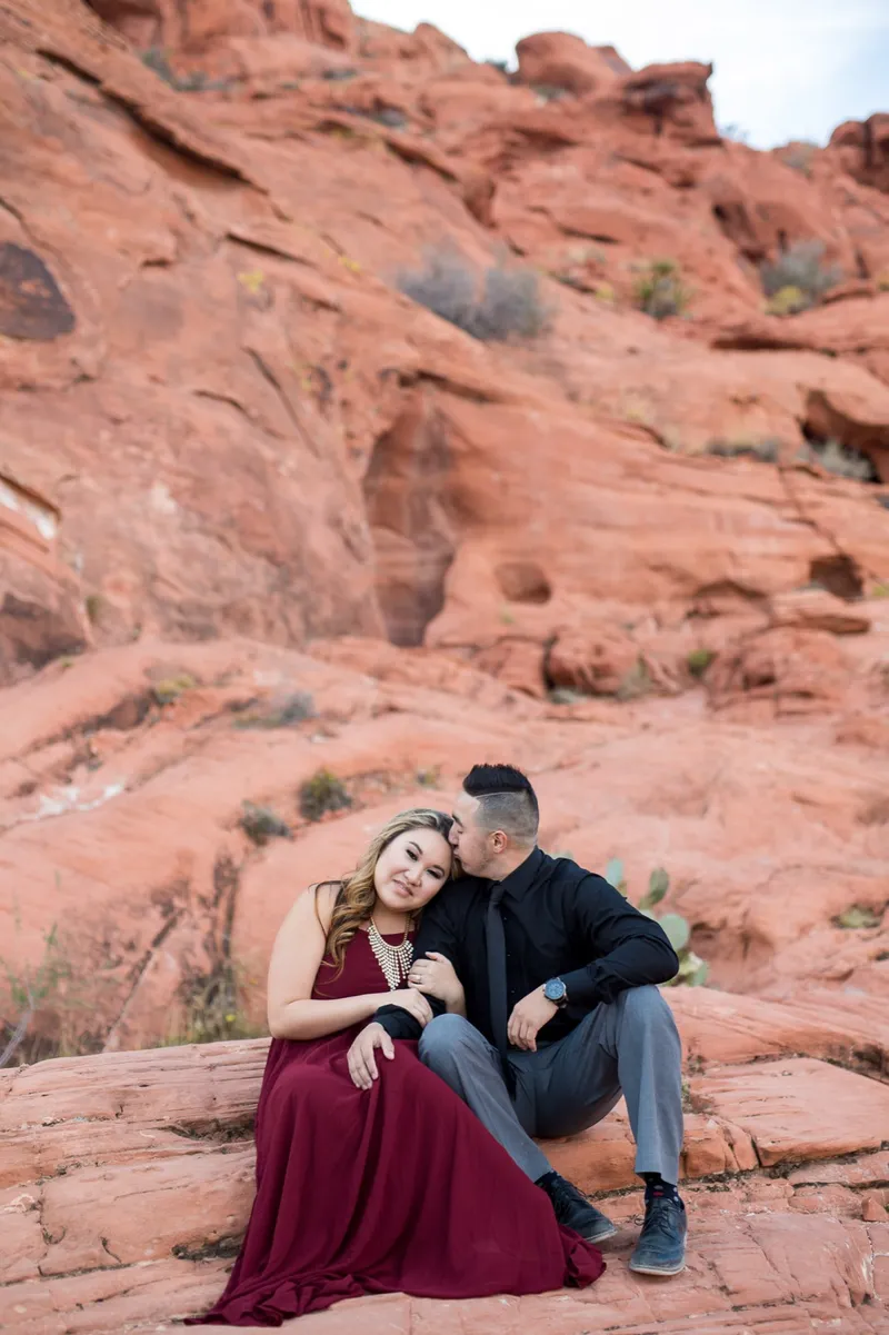 Man kissing the top of his fiancee's head as the two of them sit together on layered red sandstone