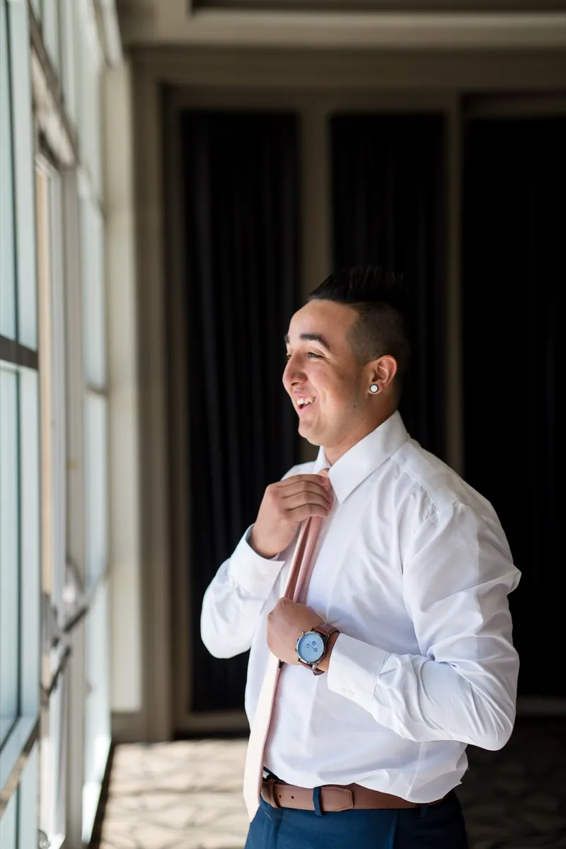 Groom in a white shirt laughing as he holds his tie in the clubhouse hallway