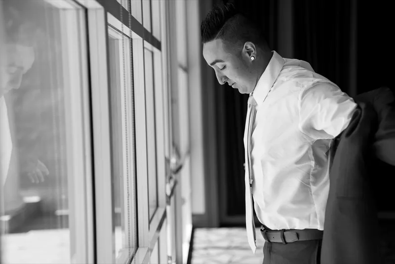Black and white photo of the groom pulling on his suit jacket beside the tall clubhouse windows
