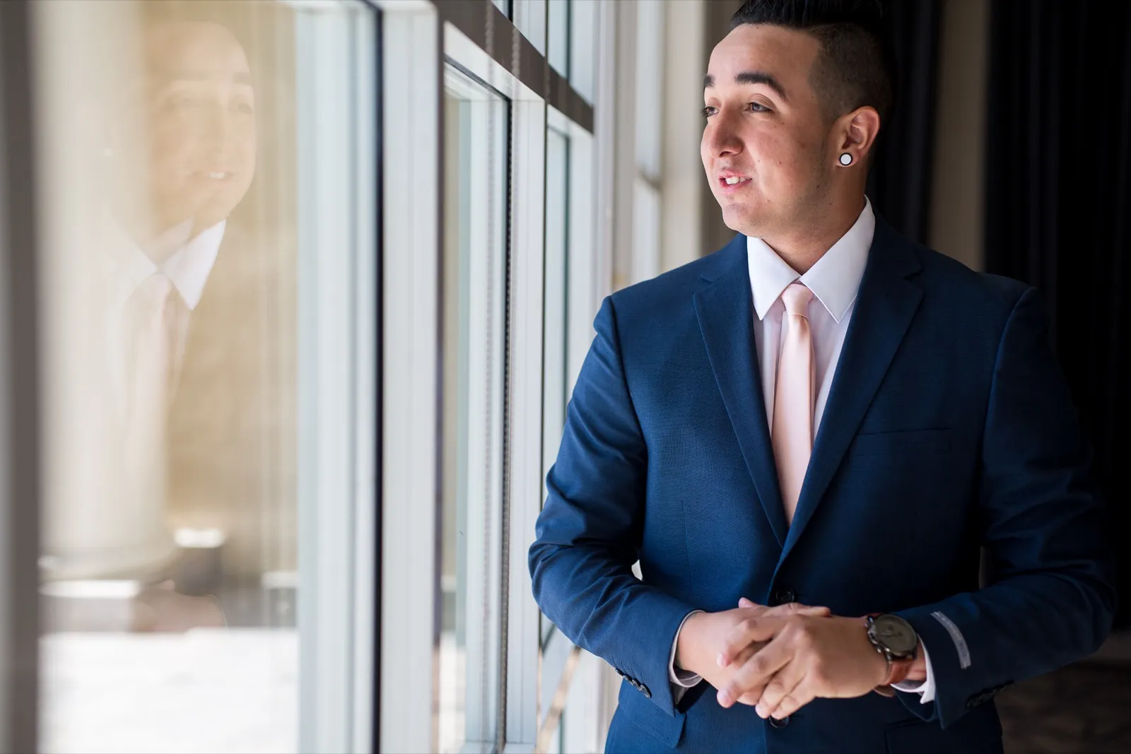Groom in a navy suit and blush tie stands at a tall clubhouse window, his reflection in the glass