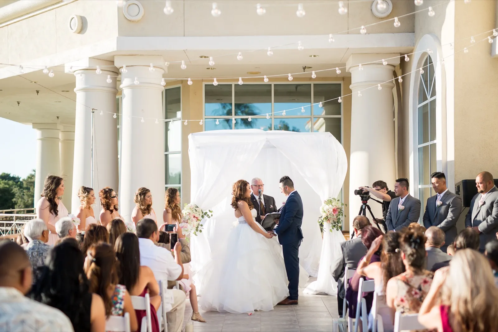 Bride and groom at the altar under the clubhouse portico, string lights overhead and guests seated on both sides