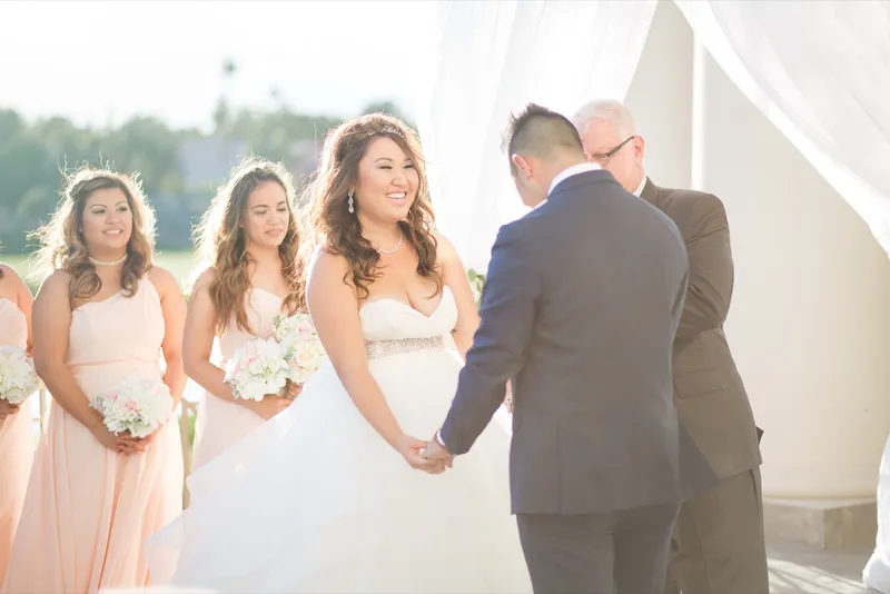 Bride in a full tulle ballgown holding both her groom's hands during the vows under white drapery hung between the columns