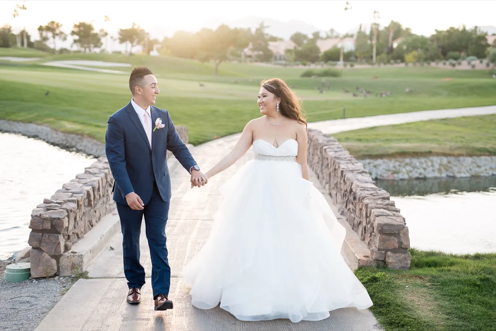 Bride and groom hold hands on a stone bridge over the water at a golf course in evening light