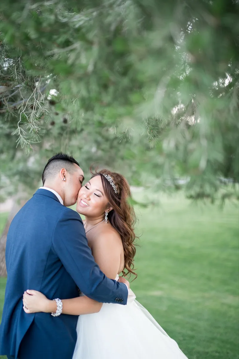 Groom kissing the bride's cheek as they hold each other under the branches of a pine on the lawn
