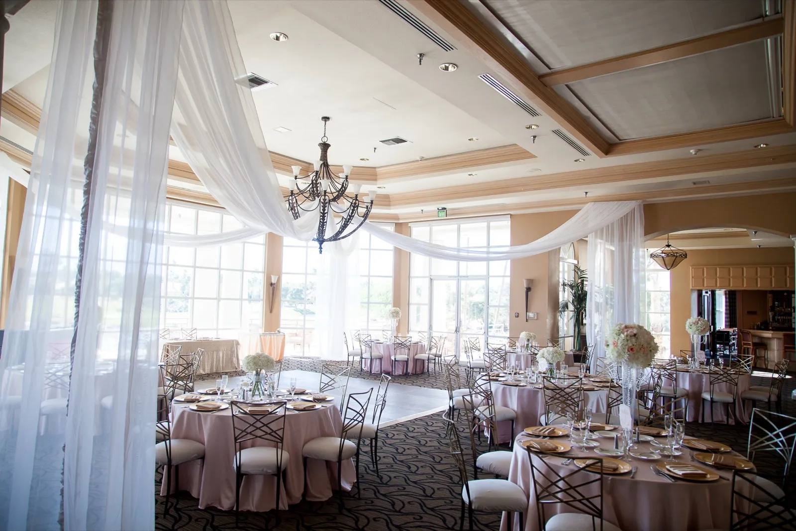Ballroom set with blush linens and gold chargers under a chandelier beside a wall of tall windows