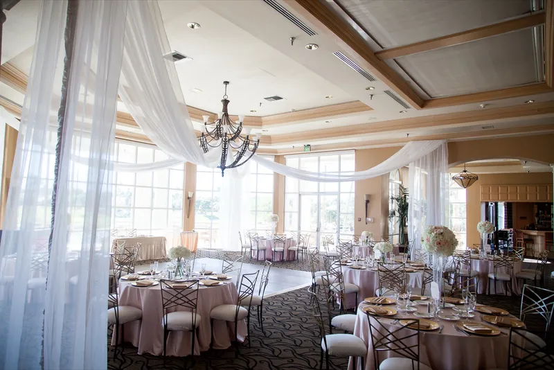 Wide view of the ballroom set for dinner in blush linens, white drapery swagged from the chandelier over the wooden dance floor and a wall of tall windows onto the course