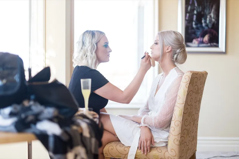 Makeup artist in black leaning in to finish the bride's makeup, the bride seated in a cream armchair by a bright window