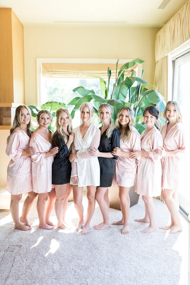 Bride and eight bridesmaids lined up barefoot in blush and black satin robes in front of tall indoor plants and a wall of windows