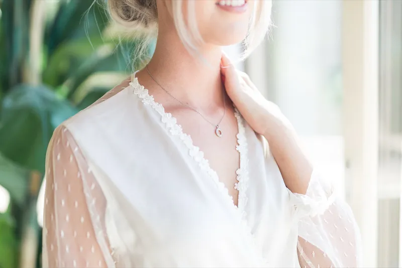 Close view of the bride's hand resting at a fine gold necklace over the collar of her lace robe
