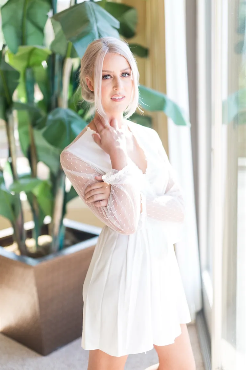 Bride in a white lace robe standing beside a tall potted banana plant, one hand at her neck, looking toward the camera