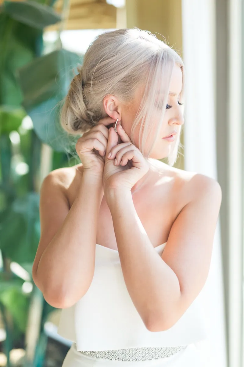 Bride already in her strapless gown, hands raised to fasten an earring, the window light coming in behind her