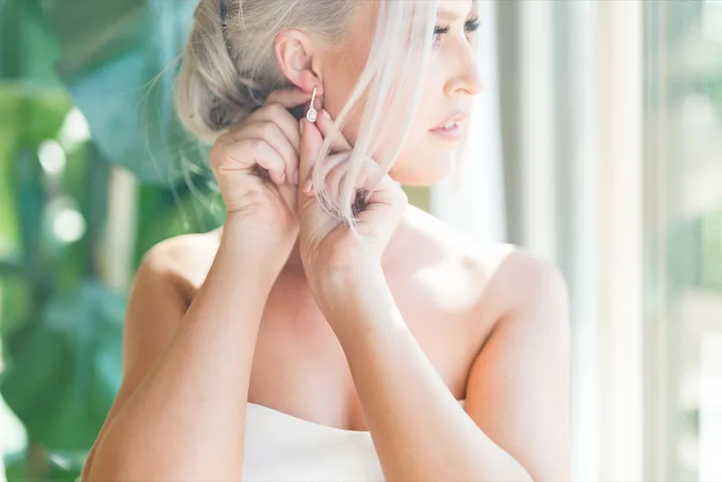 Close profile of the bride's hands putting in an earring, her hair swept into a low twist