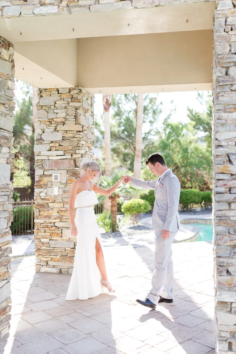 First look under a stone pavilion by the pool, the groom in a light grey suit turning around to see the bride