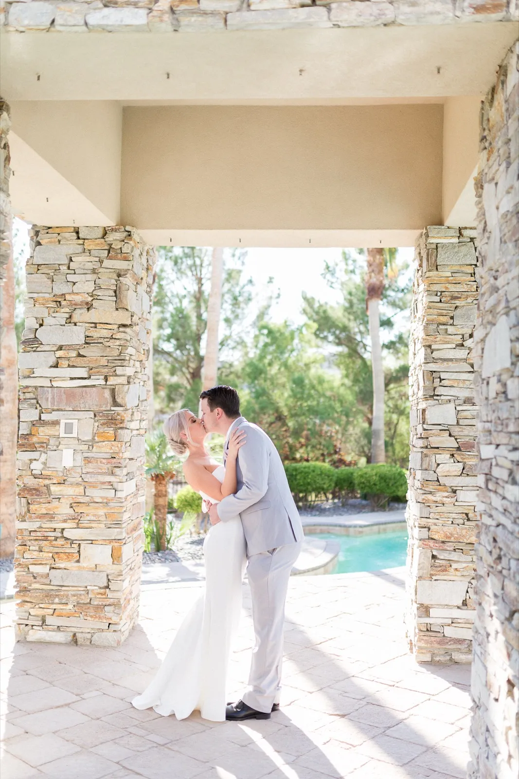 Bride and groom kiss in the shade of stacked stone columns beside a pool