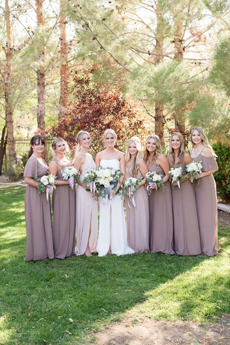 Bride with seven bridesmaids in mauve gowns on the lawn under the pines, all holding white and greenery bouquets