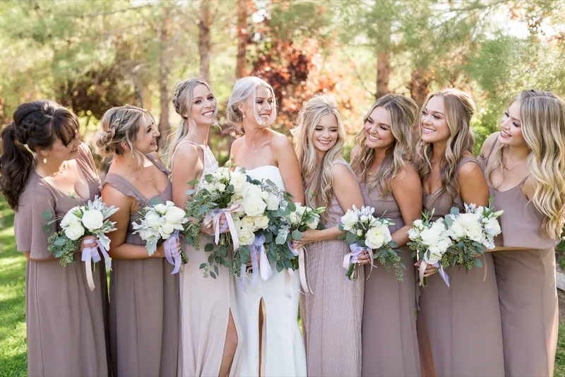 Bridesmaids leaning in and laughing around the bride on the grass under the trees