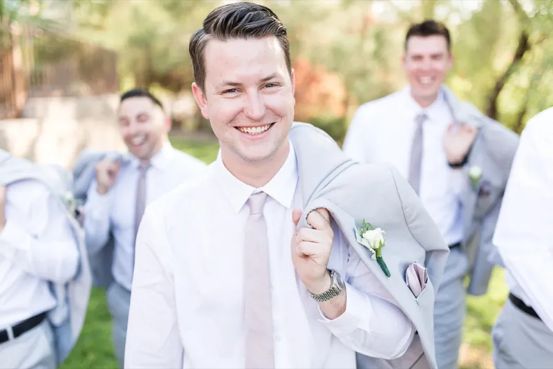 Groom grinning straight at the camera in his shirtsleeves and blush tie with his groomsmen crowded behind him