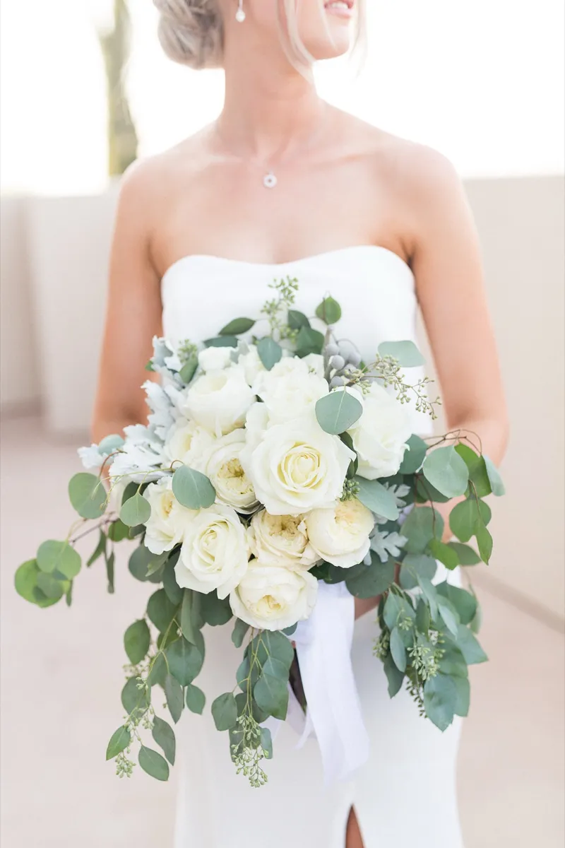 Close view of the bride holding a loose bouquet of white roses and trailing eucalyptus against her gown