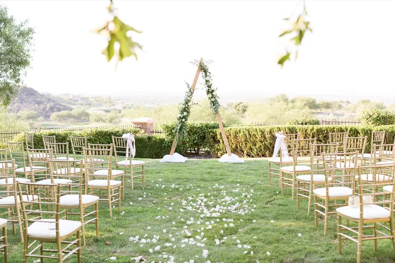 Ceremony set on a lawn with a wooden triangle arch covered in greenery and white roses, gold chiavari chairs on both sides and white petals down the aisle